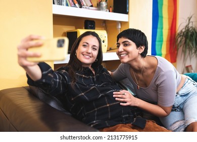 Authentic shot of happy married homosexual female gay couple with pregnant woman taking a selfie, rainbow pride flag on background - lesbian couple at home enjoying life together