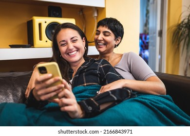 Authentic Shot Of Happy Married Homosexual Female Gay Couple Laughing And Using Mobile Phone On The Sofa For Communication And Social Media - Lesbian Couple At Home Enjoying Life Together