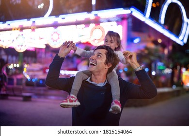 Authentic shot of a happy smiling father carrying his little daughter on a shoulders having fun together in amusement park with luna park lights at night.