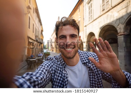 Authentic shot of young happy carefree man is making selfie or video call to a friends or parents in old city center in a sunny day. 