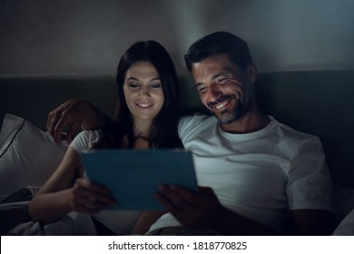 Authentic shot of young happy smiling just married couple in love is enjoying time together while using tablet for family entertainment  before asleep on a bed in a bedroom at home at night.