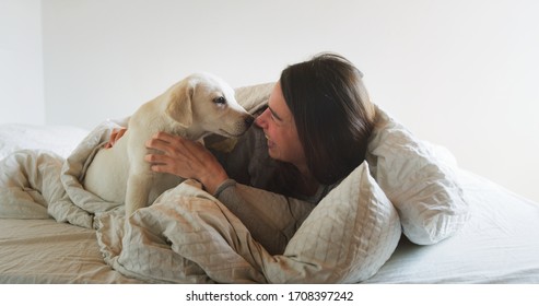 Authentic shot of an young happy woman is caressing her pedigree puppy of Labrador Retriever dog while having fun together on a bed. Concept: love for animals, friendship, authenticity, happiness,pets