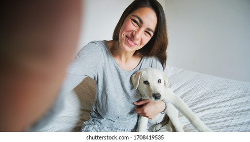 Authentic shot of an young happy woman is making a selfie or video call to a boyfriend with her puppy of Labrador Retriever dog in a bed. Concept of technology, connection,pets, family authenticity - Powered by Shutterstock