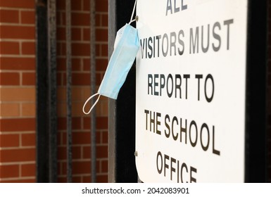 An Authentic Sign At The Entrance To A School Ground A Sign Stating Visitors Must Report To The Front Office With A Blue Protective Face Mask Hanging Off It. Blue Mask In Focus