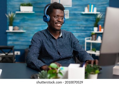 Authentic Smiling African American Man Using Laptop And Headphones To Work From Home And Learn Freelancer Webinar Or Online College Courses. Business Guy Watching Movie Indoors At Desk