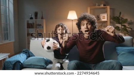 Authentic young cute asian brothers with curly hair watching soccer together, father and son reacting to victories or defeats of their team, expressing emotions - family time concept 