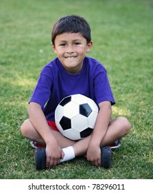 Authentic Young Latino Boy Sitting Cross-legged With Soccer Ball On Field Of Grass