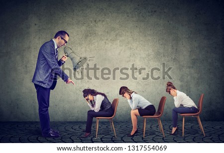 Authoritarian angry boss businessman screaming in megaphone giving orders to sad looking down female employees 