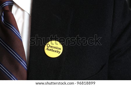 Authority Questions Authority/Close up of uncopyrighted Question Authority button pinned to black camels hair Brooks Brothers suit jacket with tie & white shirt visible.