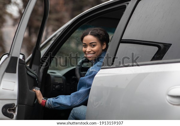 Auto blogger and test drive cars, good auto offer\
and ad. Smiling young african american lady driver sitting in car\
behind steering wheel, opens door and looks back at road in city,\
empty spaces