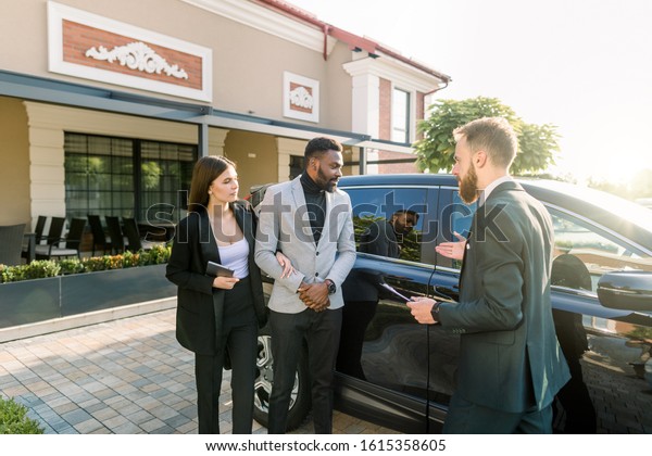 auto business, car sale, technology and people\
concept - business couple, African man and Caucasian woman with car\
dealer man standing near the black car in the yard of auto salon\
outdoors