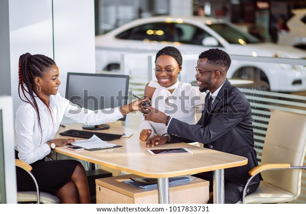 auto\
business, sale and people concept - happy african couple with\
dealer buying car in auto showroom or\
salon