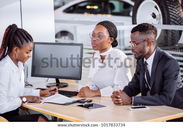 auto\
business, sale and people concept - happy african couple with\
dealer buying car in auto showroom or\
salon