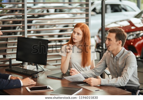 auto business, sale and\
people concept - happy caucasian couple buying car and Sales Rep\
suggests to sign a form of a Vehicle Sales Agreement at car\
dealership