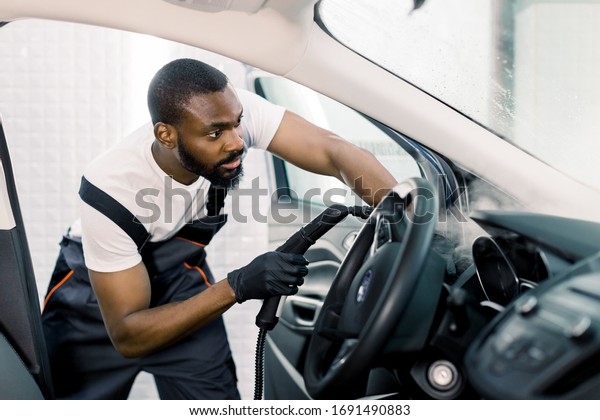 Auto cleaning service and detailing\
concept. Handsome African man in uniform cleaning interior of the\
car with hot steam cleaner. Selective\
focus.