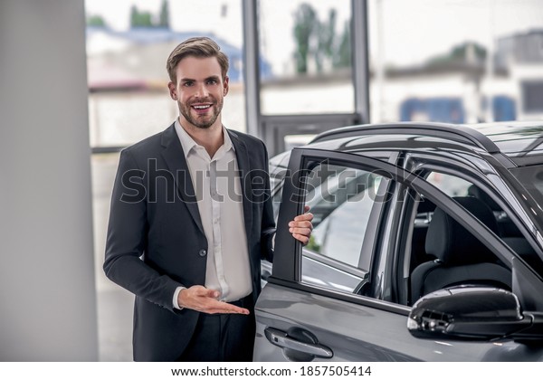 Auto dealership. Smiling\
brown-haired male presenting new car, holding car door in\
showroom