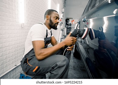 Auto detailing service, polishing of the car. Side view of young African American man worker n t-shirt and overalls, polishing blue car door with orbital polisher - Powered by Shutterstock