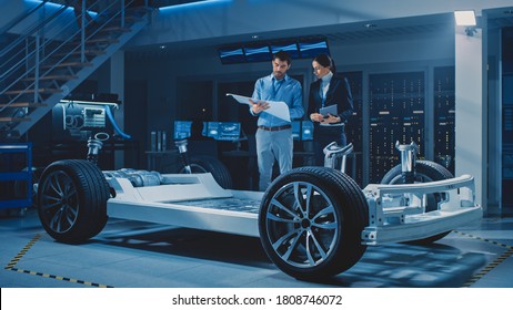 Auto Industry Design Facility: Male Chief Engineer Shows Car Blueprints Female Software Design and Integration Engineer. Electric Vehicle Platform Chassis Concept Has Wheels, Engine and Battery - Powered by Shutterstock