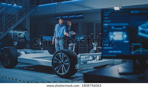 Auto Industry Design\
Facility: Male Chief Engineer Shows Car Prototype to Female Car\
Designer. Electric Vehicle Platform Chassis Concept with Wheels,\
Engine and Battery.