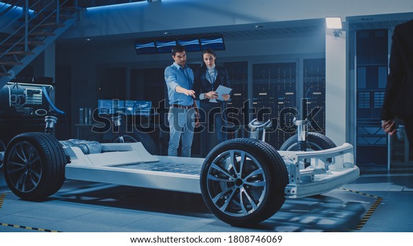Auto Industry Design\
Facility: Male Chief Engineer Shows Car Prototype to Female Car\
Designer. Electric Vehicle Platform Chassis Concept with Wheels,\
Engine and Battery.