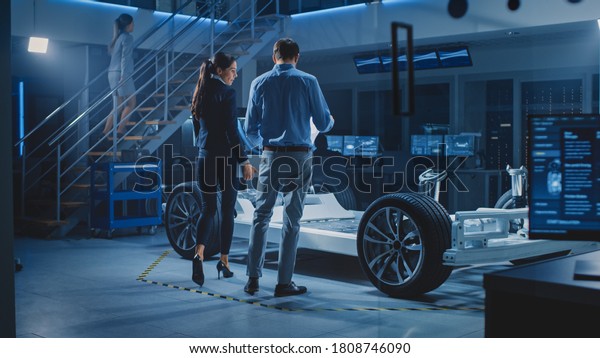 Auto Industry Design Facility: Male Chief\
Engineer Shows Car Prototype to Female Car Designer. Electric\
Vehicle Platform Chassis Concept Has Wheels, Engine and Battery.\
Screens Show 3D Cad\
Software
