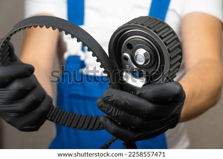 Auto mechanic in blue jumpsuit holds in his hands in black gloves roller belt tensioner of gas distribution mechanism of engine and belt. Concept of replacing belt in engine in car.