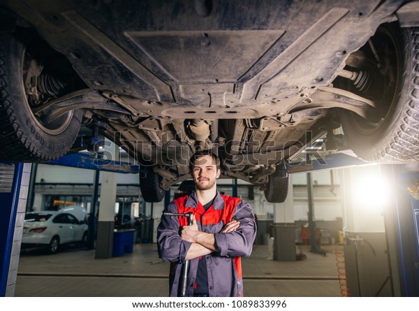 Auto mechanic crossed hands\
and looking at camera while standing under lifting car in repair\
garage