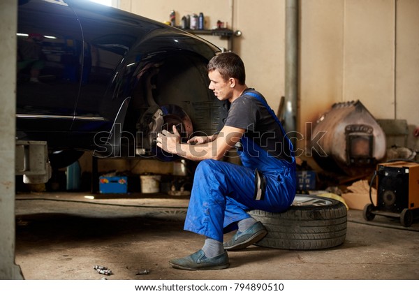 An\
auto mechanic in a dirty work uniform repairs front wheel of car in\
the garage. The guy looks carefully at the\
problem
