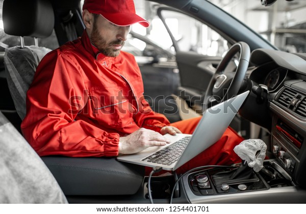 Auto mechanic in red\
uniform diagnosing car with computer sitting on the driver seat at\
the car service