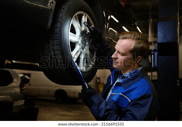 Auto mechanic in an auto repair shop making\
checklist for repairing a lifted modern car during warranty service\
and technical inspection