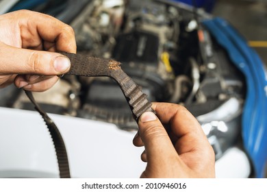 An auto mechanic shows a torn timing belt with worn teeth against the background of an open car hood close-up