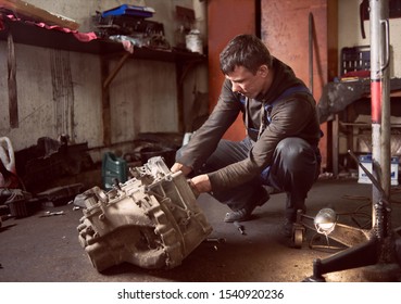 Auto Mechanic Squatting Working On Car Detail On Floor At Auto Repair Shop. Strong Repairman In Dirty Workwear In Action. Tools, Spare Parts And Garage Interior On Background