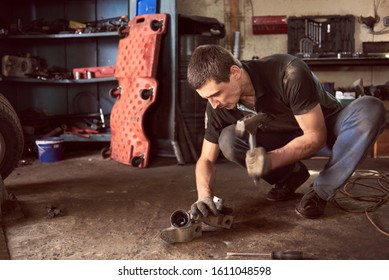 Auto Mechanic Squatting Working On Car Detail On Floor With Hammer Helping At Auto Repair Shop. Strong Repairman In Dirty Workwear In Action. Tools, Spare Parts And Garage Interior On Background