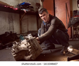 Auto Mechanic Squatting Working On Car Detail On Floor At Auto Repair Shop, Looking To The Camera. Strong Repairman In Dirty Workwear In Action. Tools, Spare Parts And Garage Interior On Background