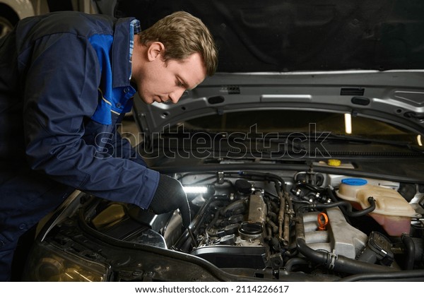 Auto mechanic, technician, car engineer with a\
flashlight lamp doing a thorough inspection for flaws in the car\
engine at car repair shop