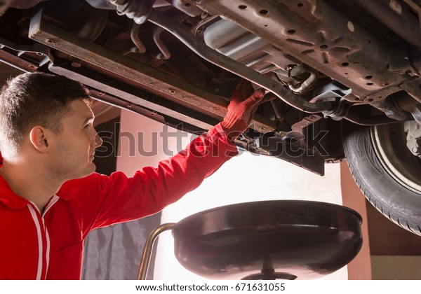 Auto mechanic in uniform working\
underneath a lifted car and changing motor oil with working\
equipment for repair and diagnostic in auto repair\
service