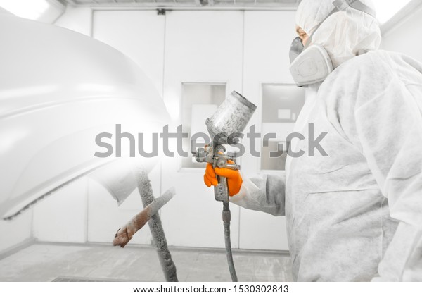 Auto mechanic worker painting\
a white car with spray gun in a paint chamber during repair\
work.