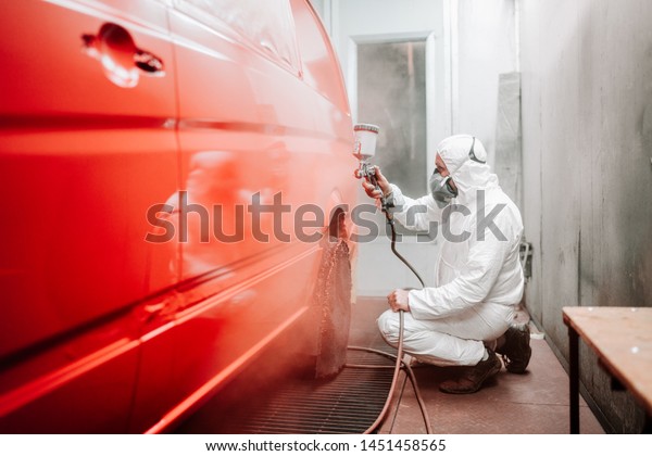 Auto mechanic working in\
automotive manufacturing industry and painting a red van in a\
special booth