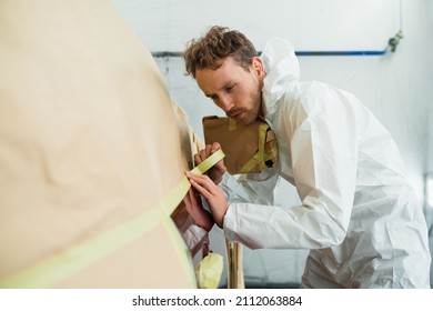 Auto painter masking car body with tape and paper before repaint. Mechanic in a white protective suit works in a garage - Powered by Shutterstock