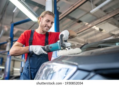 Auto repair shop. Young bearded specialist man in red tshirt and overalls improving appearance of car body in car workshop