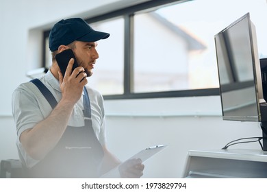 Auto Repairman Reading Data On Desktop PC While Talking With Customer Over Mobile Phone At Car Workshop. 