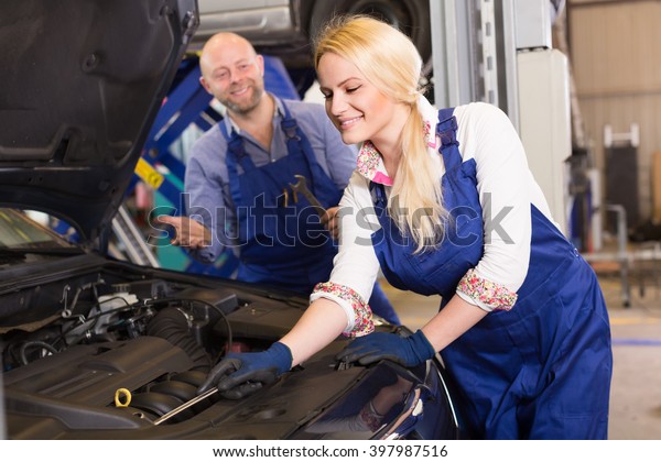 Auto service center\
crew with attractive smiling young blonde repairing a broken car at\
garage. Focus on girl