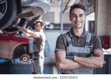 At the auto service. Handsome young auto mechanic in uniform is looking at camera and smiling while his colleague is examining car - Powered by Shutterstock