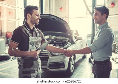 At the auto service. Handsome young auto mechanic in uniform and a client are shaking hands and smiling - Powered by Shutterstock