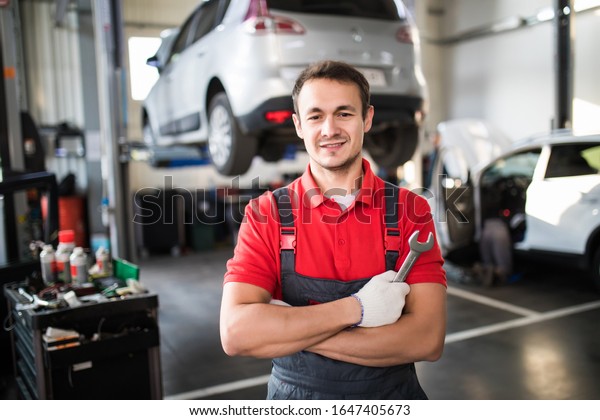 At the auto service. Handsome young auto mechanic in\
uniform is looking at camera and smiling while his colleague is\
examining car