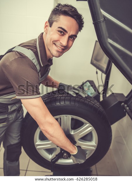 At the auto service. Handsome young auto\
mechanic in uniform is looking at camera and smiling while\
performing wheel diagnosis