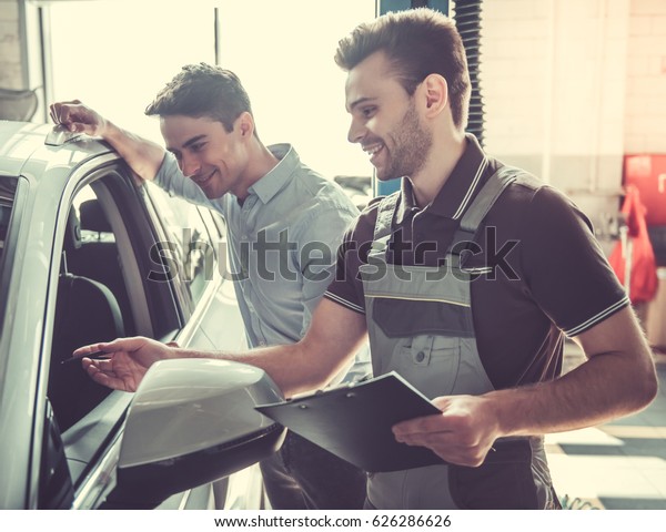 At the auto service.\
Handsome young auto mechanic in uniform is talking with a client\
and smiling