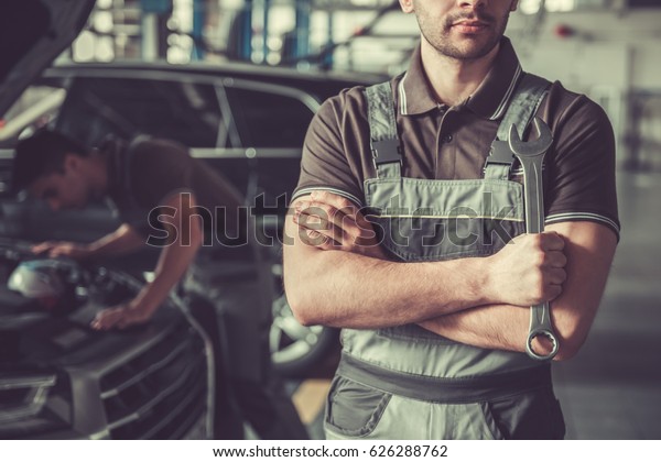 At the auto service. Handsome young auto mechanic in\
uniform is standing with spanner while his colleague is examining\
car