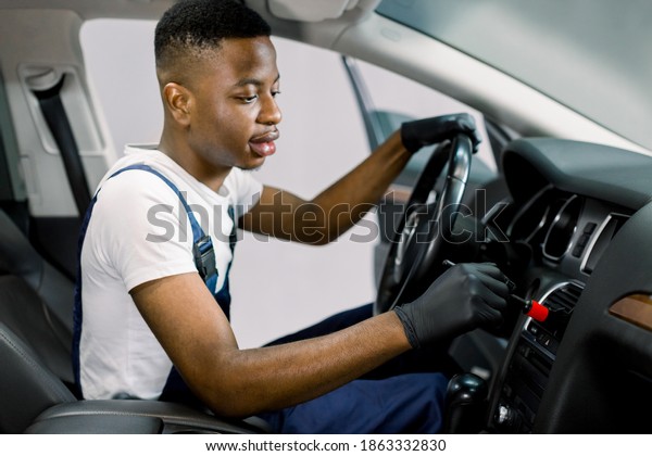 Auto service and\
professional detailing concept. Young black man worker in white\
t-shirt and overalls, makes vehicle interior cleaning, wiping front\
plastic panel with a brush