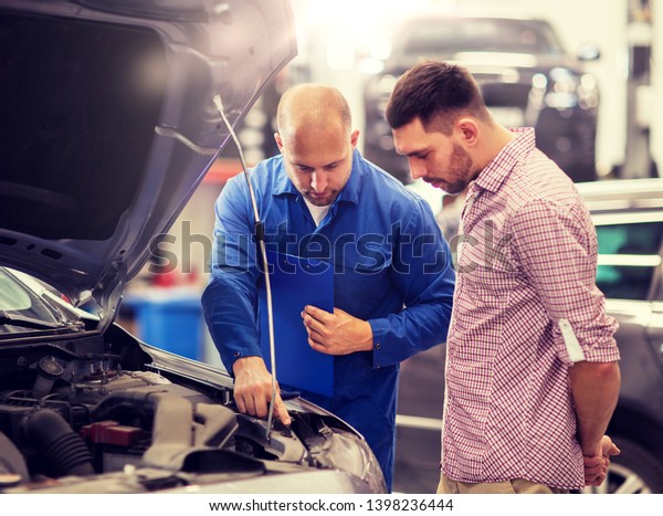 auto service, repair, maintenance and people concept\
- mechanic with clipboard and man or owner looking at broken car\
engine at shop